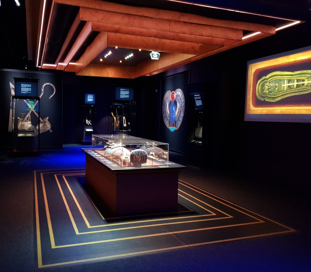 Image shows a dummy adorned with Tutankhamun's jewellery in a sarcophagus-like case. On the floor and ceiling four nested gold lines show the positions of the four nested golden shrines set around the sarcophagus in the burial chamber of Tutankhamun.