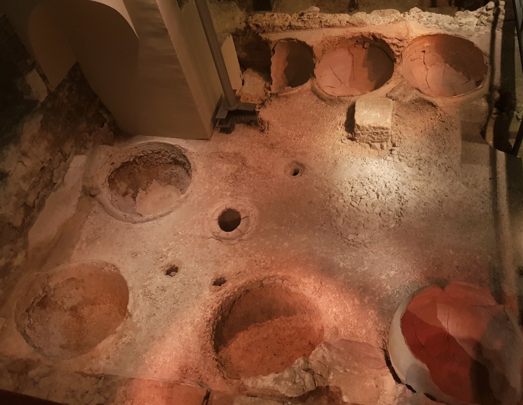 Aerial view of archaeological excavations of a courtyard with a white floor and partially sunken large pottery vessels set into it.