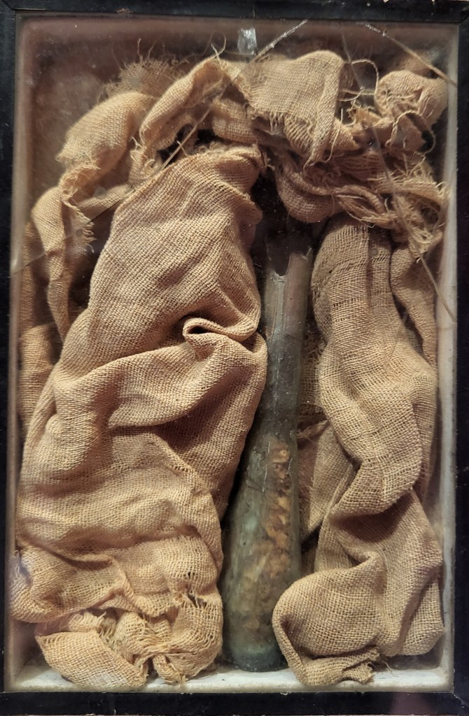 Image of a small glass vial containing brown material held in a box by browned linen.