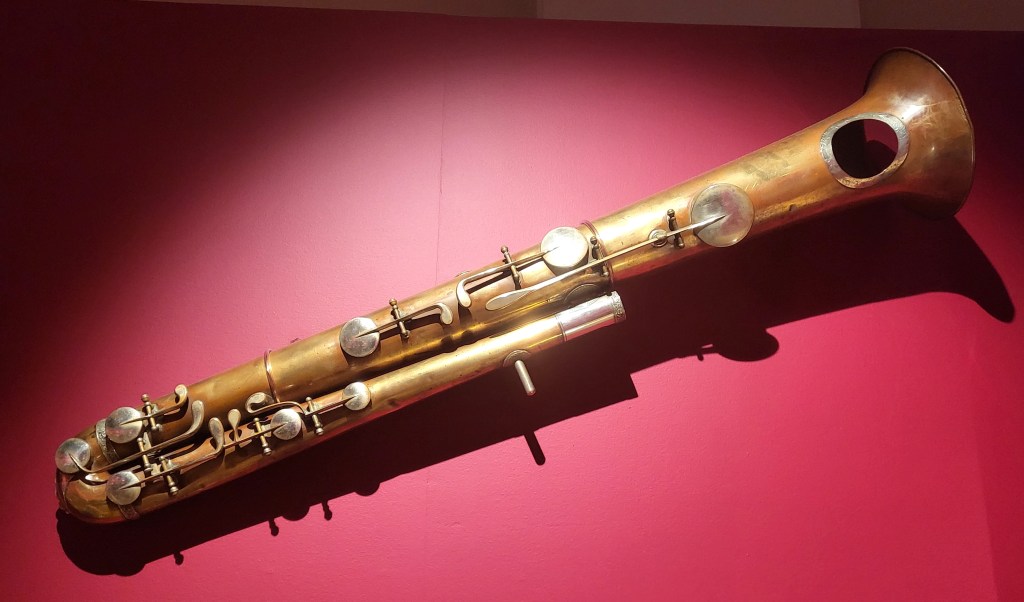 A brass musical instrument that looks partway between a tuba and a saxophone on a red background.