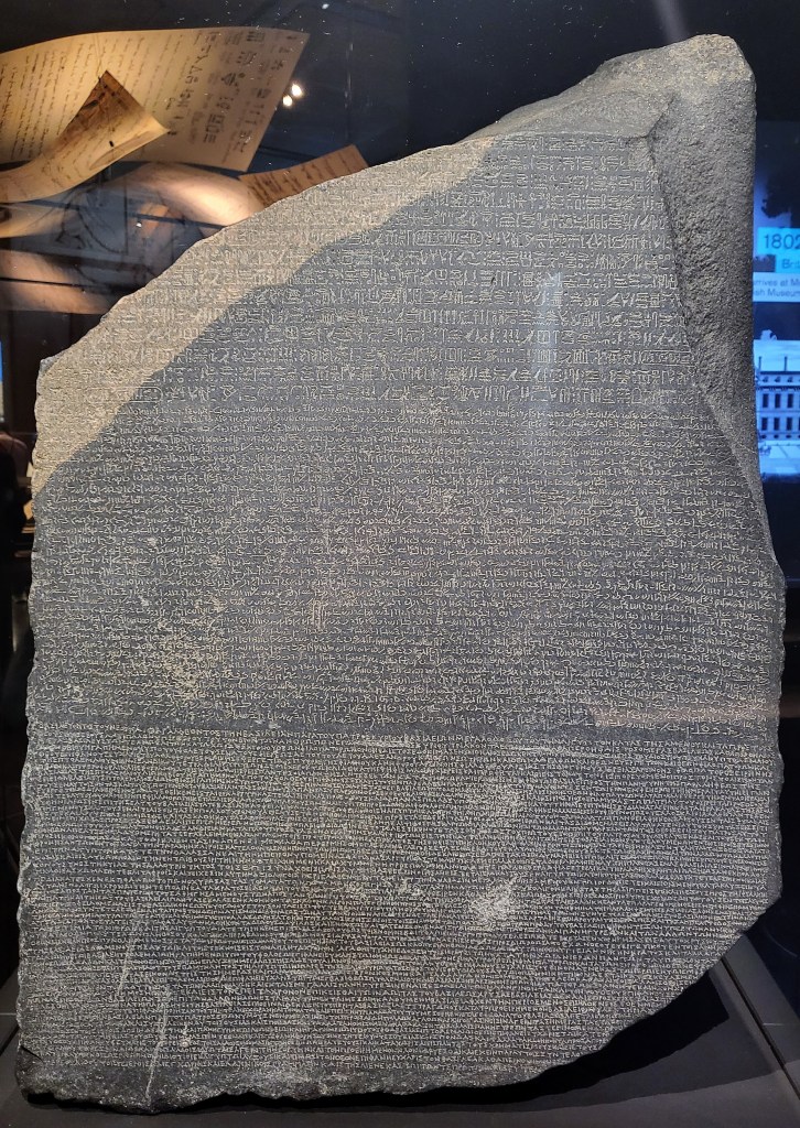 Photo of the Rosetta Stone (Hagar Rashid in Arabic) - a large lump of blue black stone irregularly broken and divided into 3 sections. The top hieroglyphs, the middle demotic and the bottom Greek.