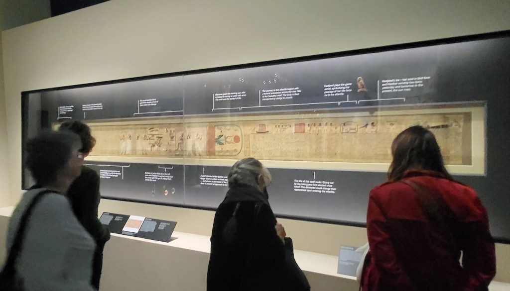 Photo from the British Museum exhibition showing visitors in front of a display showing the decorated funeral papyrus of Nedjmet. Around the papyrus the black case has information describing what the different sections mean.