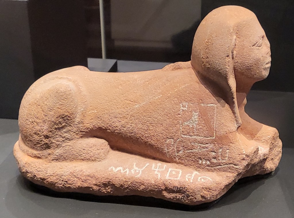 Photo of a small pink sandstone Sphinx with hieroglyphics on the shoulder and proto-Sinaitic writing on the
base