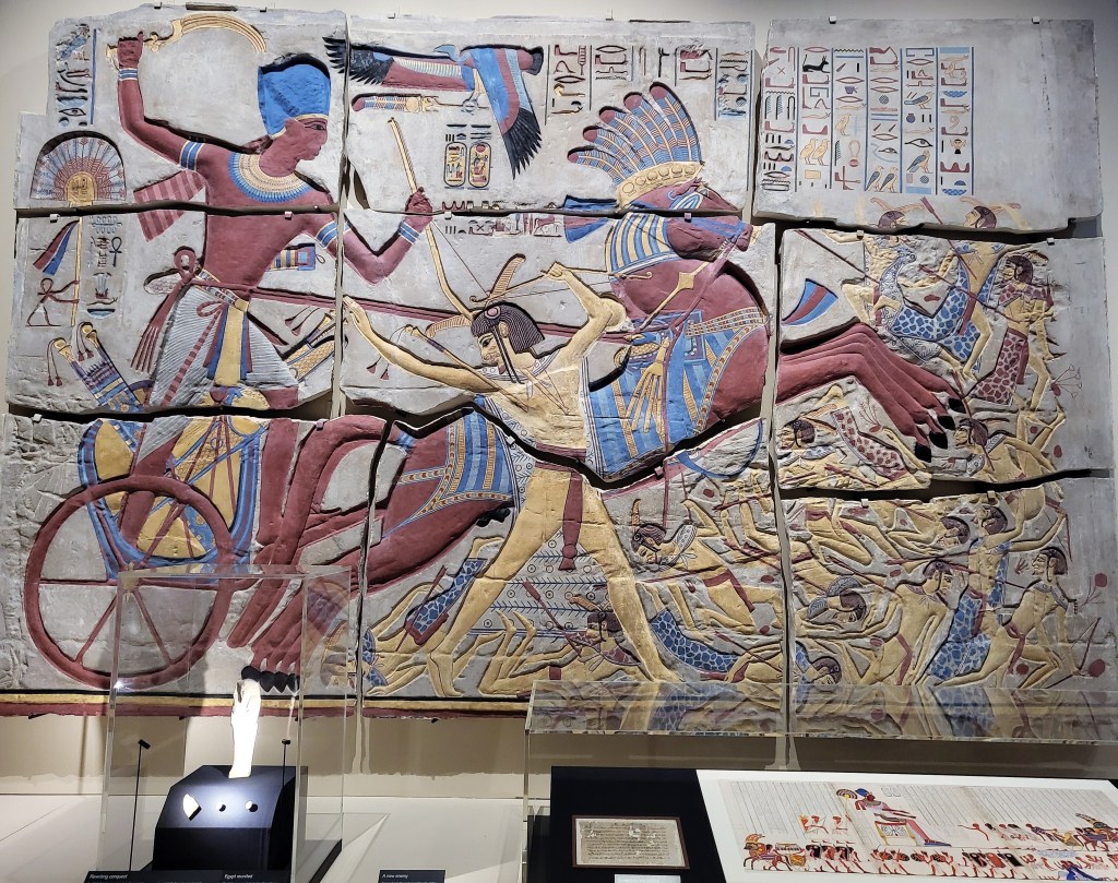 Photo of the coloured plaster cast of a relief showing Pharaoh Seti I in a chariot smiting a Libyan with a khepesh sword and riding over others.