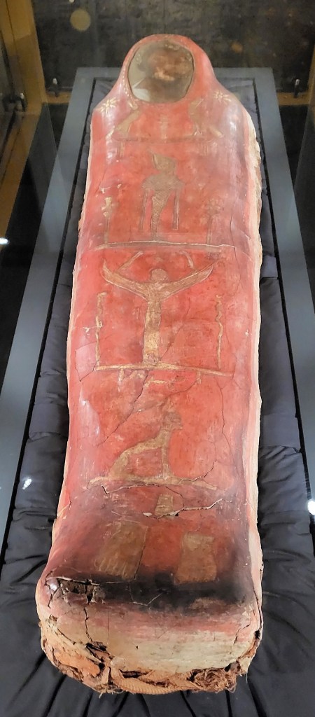 Picture of a mummified ancient Egyptian with a portrait face panel over his head and gilded Egyptian deities on a red background down the torso and legs.