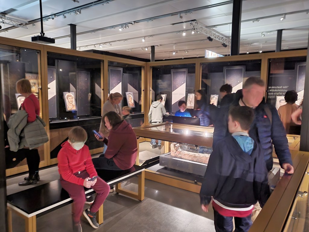 Photograph of a busy exhibition room with upright cases containing individual Faiyum portraits. In the centre is a place to sit and a case containing a Faiyum portrait in situ on the original mummified person. 