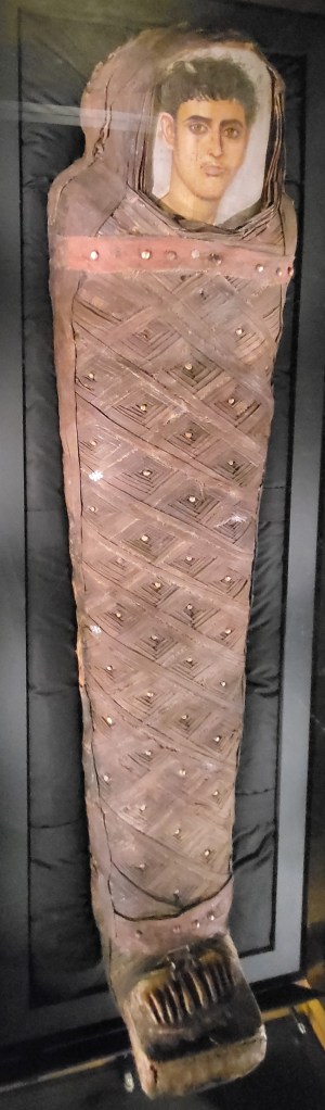 Photo of a completed mummified young man with a portrait panel covering his face. He has a gilded foot case and decorative bandaging of lozenges, each with a gold bead at the centre.