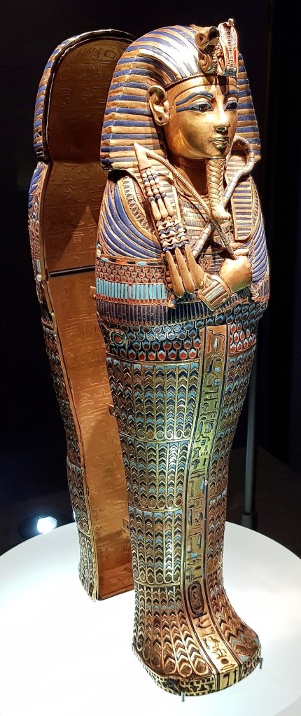 Photo of a miniature mummiform coffin of Tutankhamun, gilded and inlaid with precious stones, shown standing up in a display case. 