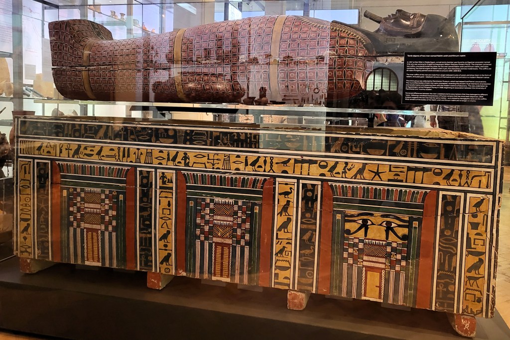 Photo of a rectangular Middle Kingdom coffin decorated with false doors and hieroglyphs below and anthropoid coffin decorated with a blag wig and red lozenge decoration across the body. 