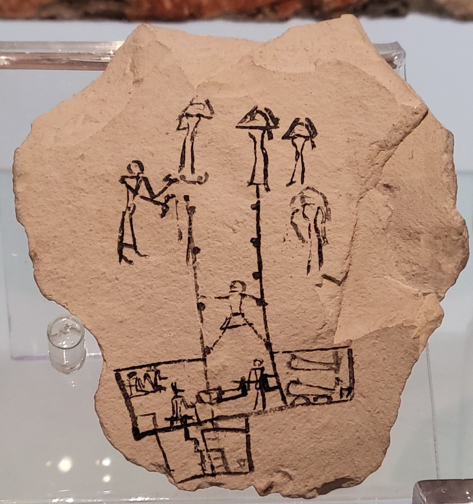 Photo of a limestone ostracon showing a pen a ink scene of a burial. Mourners are shown at the top of a vertical shaft with hand and foot holds. One person descends the shaft. At the bottom another person moves the sahw into position amongst two others. A person wearing an Anubis mask stands by to guide the sahw into position in a lower chamber accessed by stairs. 