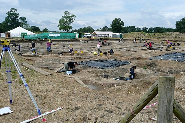 An open excavation trench, with various large tented structures beyond. The trench is filled with various archaeologists working on the features within it. Some features are covered with tarps. A spoil heap in the distance is old enough to have grassed over. 