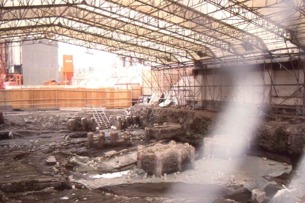 A photo of a wet trench with surviving pile foundations cutting through archaeological remains, covered by a temporary roof. 