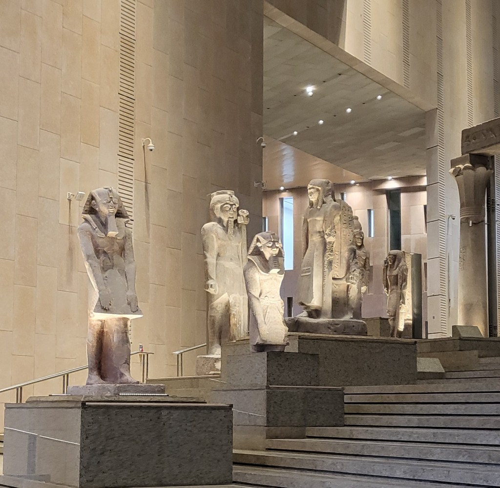 Photo of the royal statues on the Grand Staircase showing Hatshepsut next to a statue of Thutmose III, usurped from her. Hatshepsut is on a lower step but has a large plinth that raises the top of the headress above that of Thutmose III.