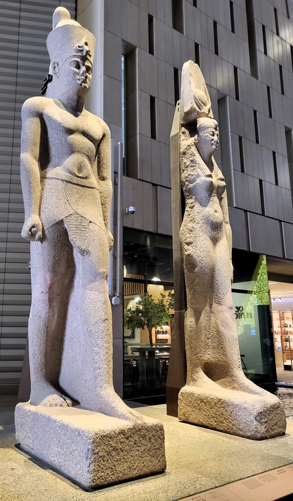 Photo of two over life-size, red granite statues showing a Pharaoh and Queen of the Ptolemaic period in Pharaonic dress. Cafes and shops are visible behind them.