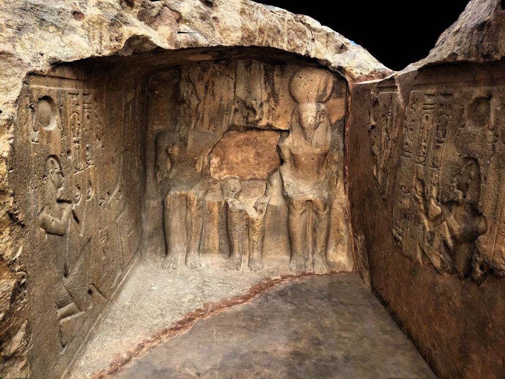 Photo of the interior of a rock-cut naos, with inscriptions on the walls and three divine statues carved out of the rear wall of the barrel-vaulted interior.