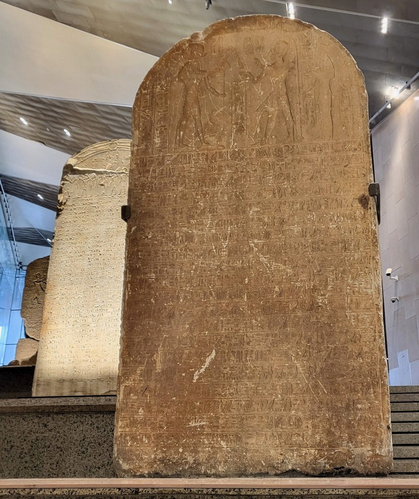 Photo of a quartzite stela with an image of Pharaoh before a falcon-headed Re, above rows of hieroglyphic text. Parts of two further stelae are visible behind.