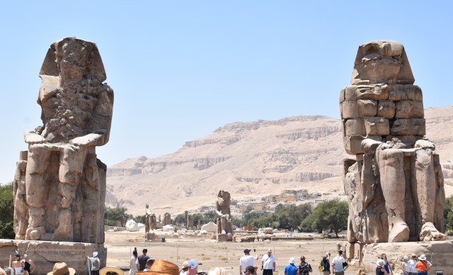 Photograph of two colossal ancient Egyptian seated statues with other partially excavated and erected statues, stelae and columns behind.