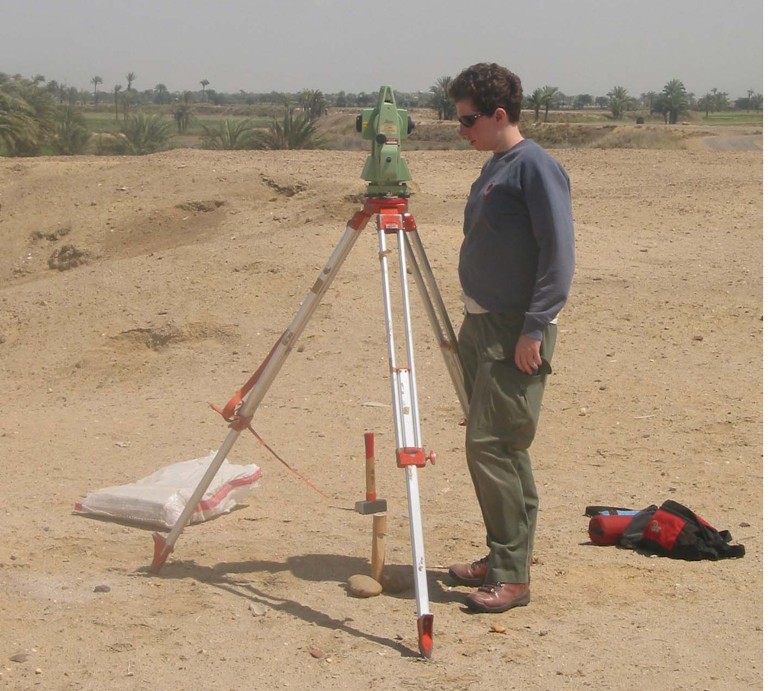 Photograph of a woman looking into a total station that has been set up over a wooden grid peg in a desert area.
