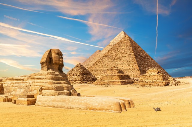 An AI generated image showing the three Giza pyramids lined up behind the pyramid of Menkaure. In front of the pyramid of Menkaure are three Queen's pyramids,and in front of them a ludicrously sized Great Sphinx, with a diminuative camel in front of him.