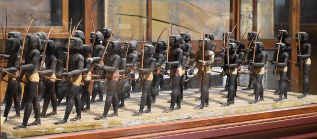 A museum case containing a group of carved wooden bowmen wearing short white and red kilts and carring self bows. 