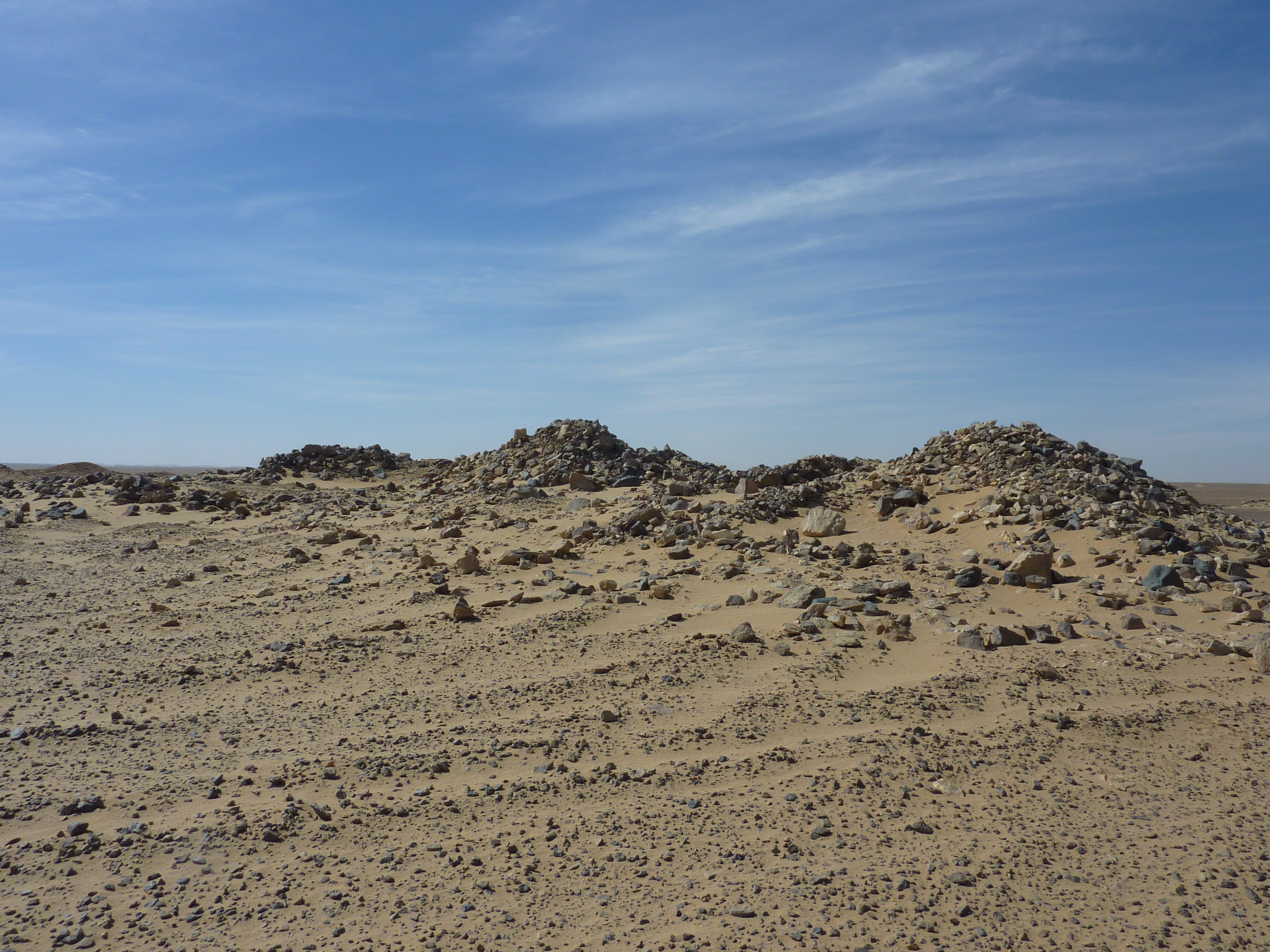 Photograph of 3 mounds of stone, variously denuded with tracks running up to them. The cairn in the right foreground is best preserved. The cairn in the rear left is badly denuded.