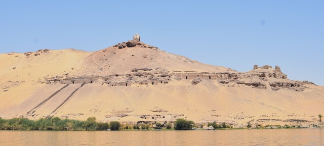 Photograph showing a steeply sloping escarpment above the Nile, with a domed structure on the top. Halfway up are two terraces with two rows of tomb entrances. At the left end of the row two causeways lead from tomb entrances down to the Nile. 