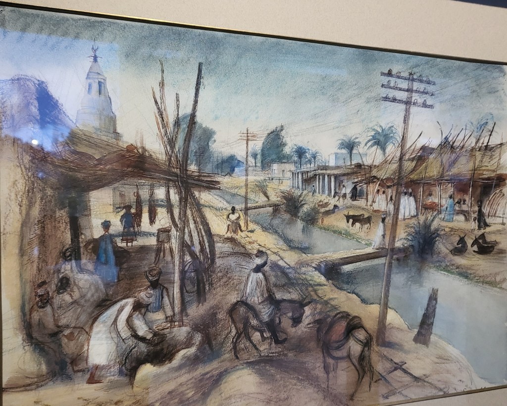 Painting of a Nubian village, with buildings on either side of a canal bridged by half palm trees. On the centre right various people go about their business around buildings beside the canal. In the left foreground is a building with a lead-to attached to its front where people sit. Two hanging shapes at the back of the lean-to might be meat awaiting purchasers. In the centre foreground a man rides a donkey and donkeys are also visible on the other side of the canal. The left rear of the picture shows a mudbrick tower typical of traditional Nubian mosque minarets.