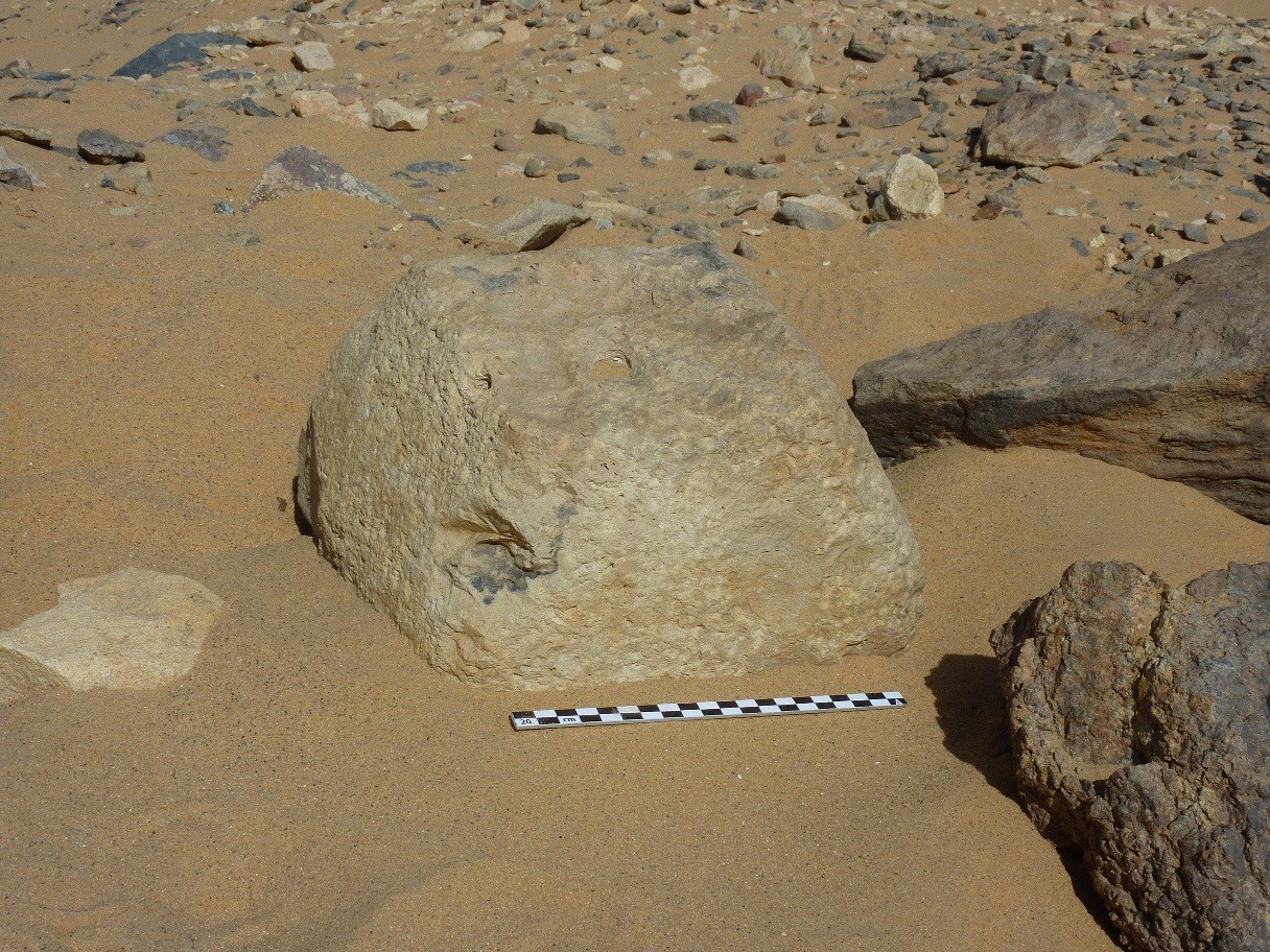 Photograph of a small stone, roughly shaped into a pyramid form sat upright on the sand.  A scale suggests it is roughly 25-30cm long.